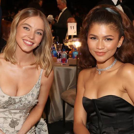 Zendaya y Sydney Sweeney durante el rodaje de Euphoria, de pie en un set con luces cálidas y cámaras alrededor; ambas lucen concentradas pero con gestos serios, reflejando la tensión entre sus personajes y los rumores de distanciamiento tras bambalinas.